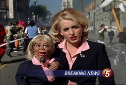 A blonde woman in a black blazer and pink shirt holds a small doll dressed in matching attire.