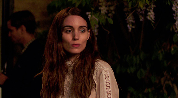 A woman with long wavy reddish-brown hair wearing a white lace blouse looks to the side at night.
