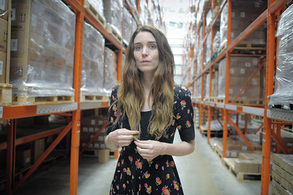 A woman in a floral dress stands in a warehouse aisle between tall orange storage racks.