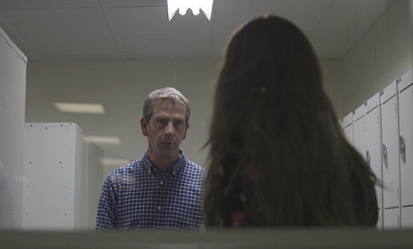 A man in a blue checkered shirt looks toward a woman with long dark hair in a hallway with white lockers.