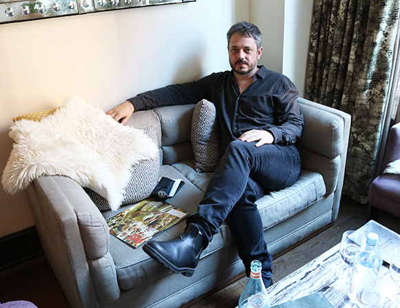 A man with dark hair and a beard sitting on a grey sofa wearing a black shirt and dark pants.