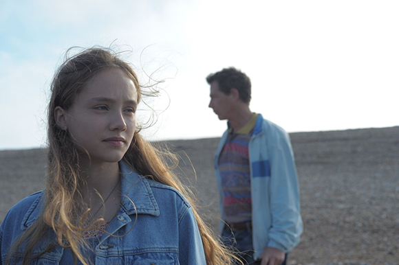 A young woman with long blonde hair in a denim jacket looks off-camera against a bright, open landscape.