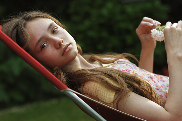 A young woman with long brown hair lies in a red hammock, looking at the camera while holding white flowers.