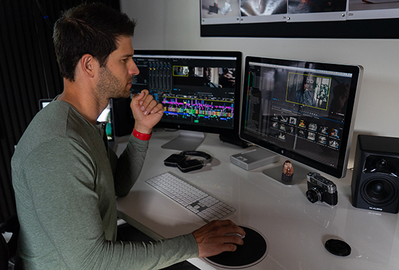 A man sits at a white desk using two computer monitors to edit video content.