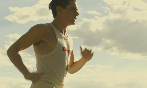A man in a white tank top runs outdoors against a bright, cloudy sky.