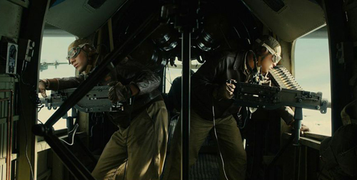 Two people in flight gear operating large machine guns inside the cargo hold of an aircraft.