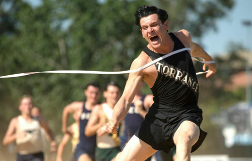 A male runner in a black tank top crosses a white finish line tape with an intense expression.