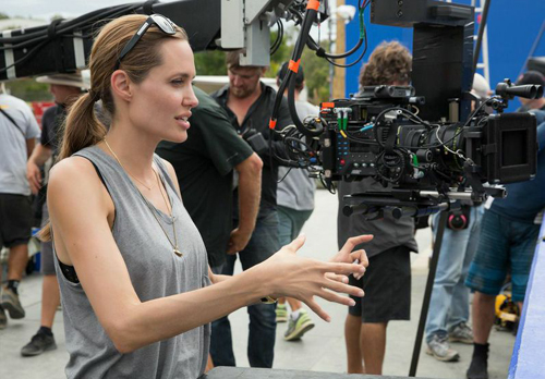 A woman in a grey tank top gestures with her hands near a large professional film camera on a set.