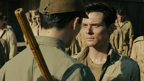A man in a tan military uniform looks intensely at another man wearing a cap.