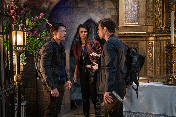 Three people stand in a stone room with gold decor, engaged in a serious conversation.