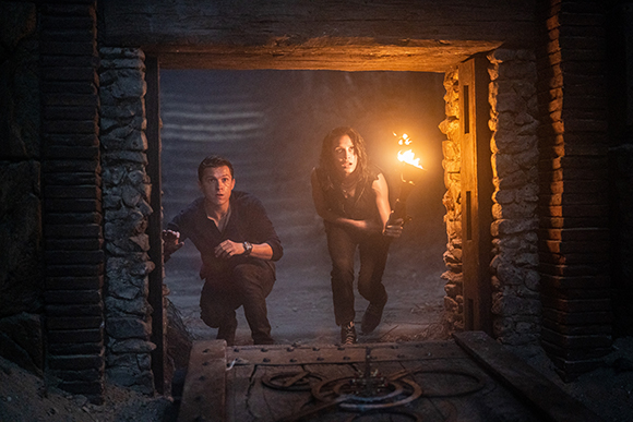 Two men crouch in a dark stone doorway, one holding a lit torch in a dim tunnel.