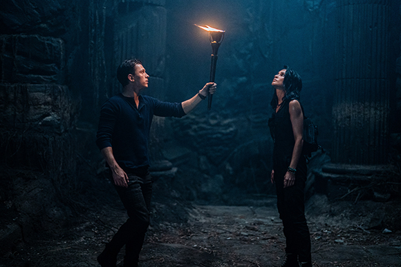 A man holds a flaming torch up toward a woman in a dark, rocky cave.