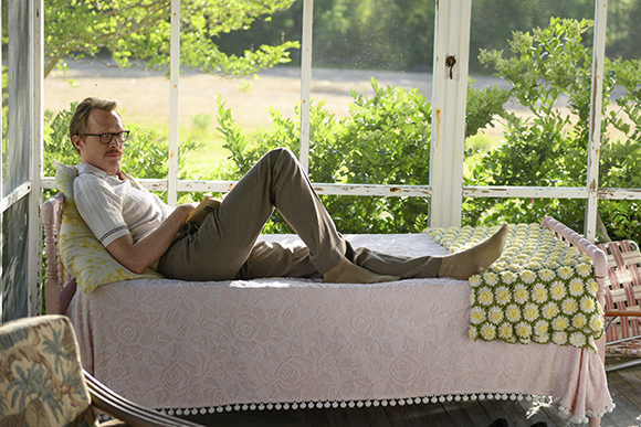 A man with glasses lies on a bed in a screened porch, reading a book amidst green foliage.