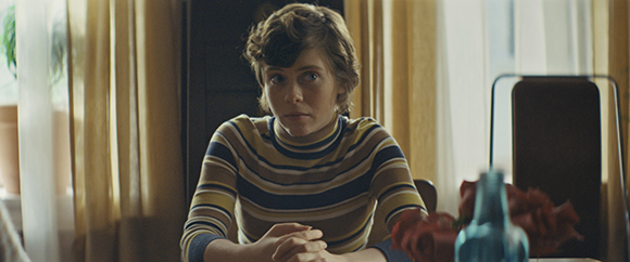A woman with short wavy hair wearing a yellow and blue striped turtleneck sits at a table.