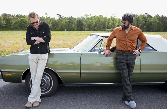 Two men lean against a vintage green convertible parked on a road near a grassy field and trees.