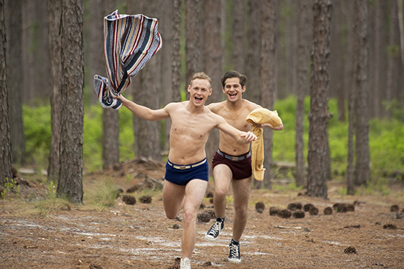 Two shirtless men running through a pine forest wearing briefs and holding towels.