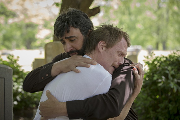 Two men sharing an emotional hug outdoors near gravestones and greenery.