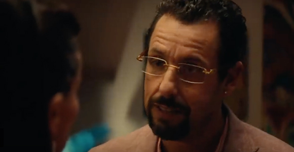 A man with dark hair, a goatee, and gold-rimmed glasses looks toward a person out of focus.