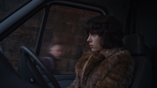 A woman with dark hair wearing a brown fur coat looks out a car window at night.