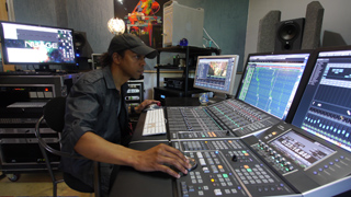 A man in a cap operates a large digital audio mixing console in a recording studio with multiple monitors.