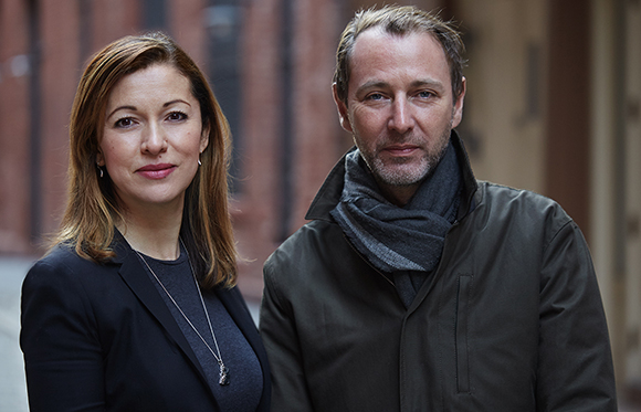 A woman in a black blazer and a man in a dark jacket and grey scarf standing together outdoors.