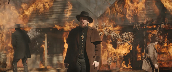 A man in a dark coat and wide-brimmed hat stands in front of wooden buildings engulfed in flames.