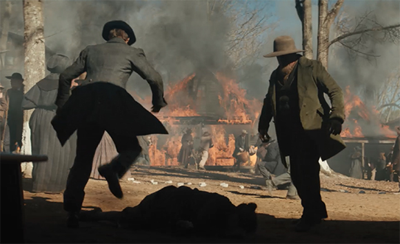 People in period clothing run through a clearing near burning buildings and smoke.