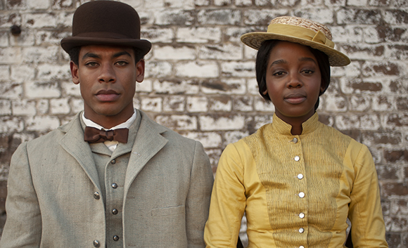 A man in a bowler hat and grey suit stands next to a woman in a yellow dress and straw hat.