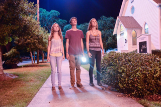 Three teenagers stand on a sidewalk at night, looking up toward the sky near a small white church.