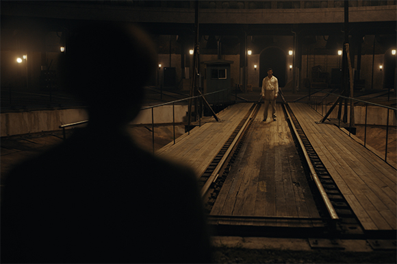 A person stands on a wooden platform under dim lights in a large, dark industrial space.