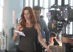 A woman with long red hair reads a paper while a man and a large film camera stand behind her.