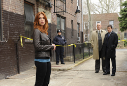 A woman in a brown leather jacket stands in the foreground near yellow police tape and a brick building.