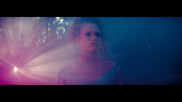 A woman with curly hair is lit by dramatic pink and blue light in a dark, misty environment.