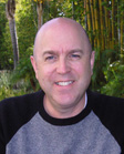 A smiling bald man wearing a grey and black sweatshirt outdoors with trees in the background.