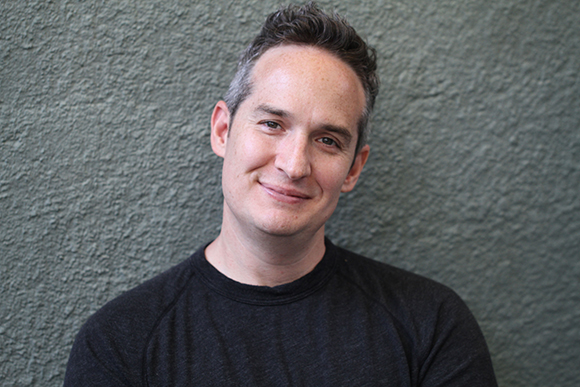 A man with short dark hair and a slight smile wearing a black long-sleeved shirt against a gray wall.