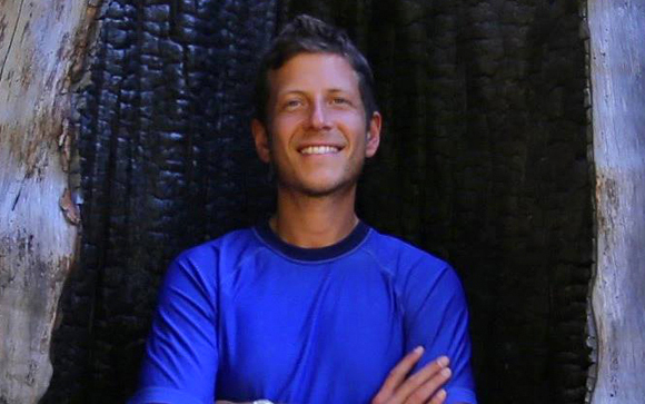 A smiling man wearing a blue shirt stands in front of a dark, charred tree trunk.