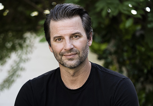 A man with dark hair and a salt-and-pepper beard wearing a black t-shirt, smiling outdoors.