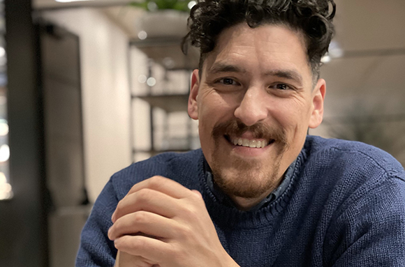 A smiling man with dark curly hair and a goatee wearing a blue knit sweater.