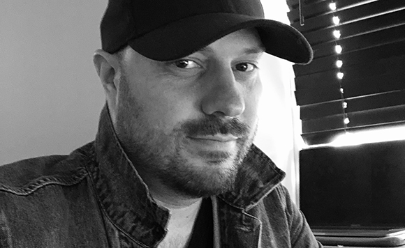 Black and white close-up of a man with a beard and mustache wearing a dark baseball cap and denim jacket.