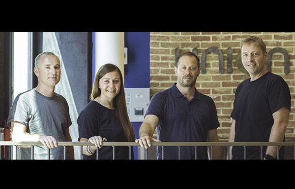 Four smiling adults stand behind a metal railing in front of a brick wall and blue pillar.