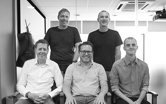 Five men posing for a group portrait in an office, three standing and two seated.