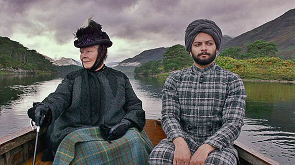 An elderly woman in black and a man in a grey turban and plaid suit sit in a wooden boat on a calm lake.