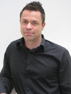 A man with short dark hair wearing a black button-down shirt against a white background.