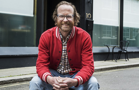 A man with glasses and a beard wearing a red bomber jacket sits outdoors in front of a dark building.