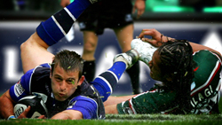 A rugby player in a blue jersey dives toward the ground with the ball while being tackled by an opponent.
