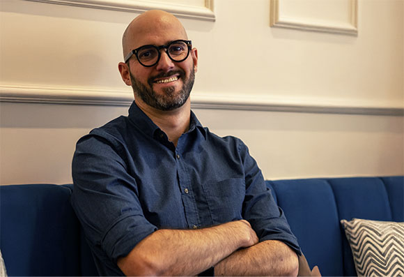 A bald man with glasses and a beard smiles while sitting on a blue couch with his arms crossed.
