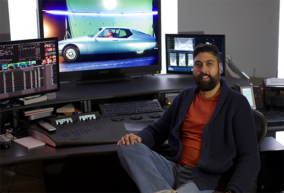 A man with a beard sits at a desk with three monitors and a video editing console.