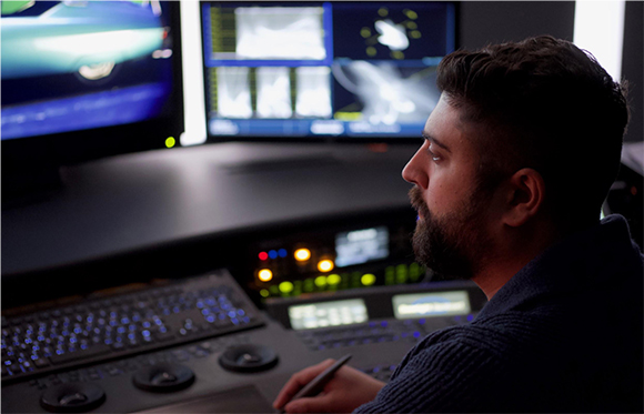 A man in profile sits before dual computer monitors and a professional video editing control surface.