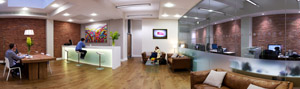 A spacious office lounge with wooden floors, a white reception desk, leather sofas, and glass-walled meeting rooms.