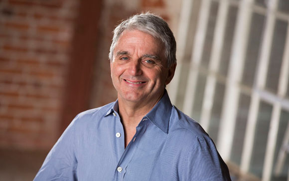 A smiling middle-aged man with gray hair wearing a blue button-down shirt.